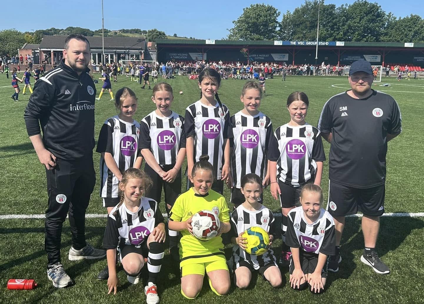 Huncoat Under 10s Girls football team in their LPK-sponsored striped kit with coaches at a tournament.